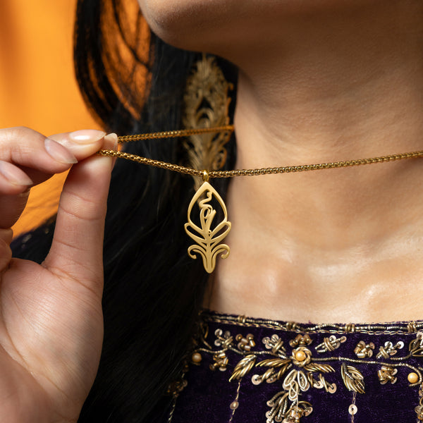 A delicate gold-plated chain necklace featuring a stylized, teardrop-shaped pendant with intricate cut-out details. The necklace is worn by a person with dark hair and is shown against a blurred orange background. A hand with henna designs is gently touching the pendant.