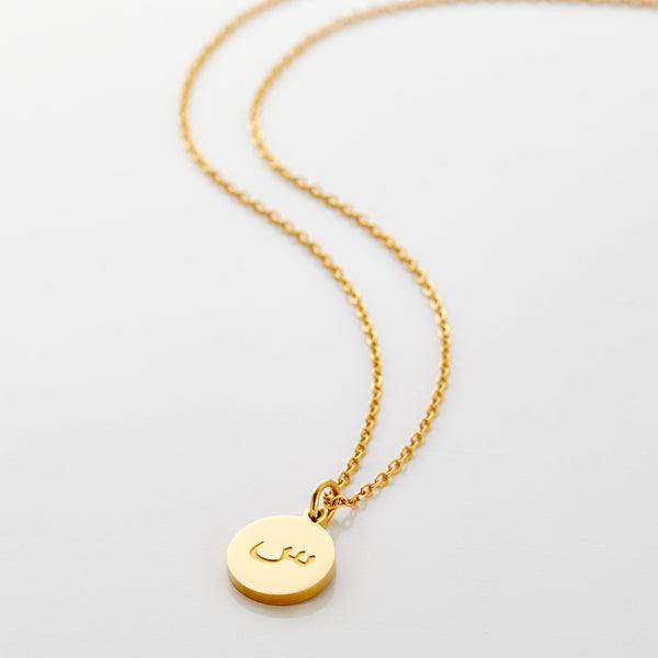 A delicate gold-plated necklace with a circular pendant. An Arabic letter is elegantly engraved in the center of the pendant. The necklace has a thin chain and is displayed on a light beige background. The specific Arabic letter is clearly visible within the circular frame of the pendant.
