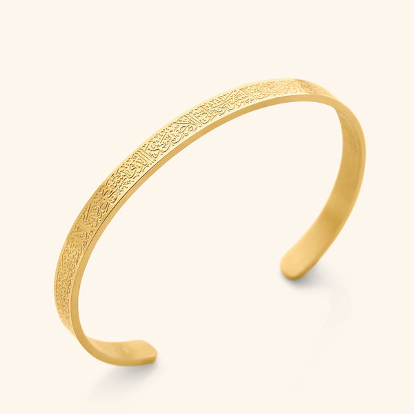 A close-up shot showcases a slender, open-ended gold cuff bracelet. The bracelet's surface is intricately engraved with Arabic script, identified as the Ayatul Kursi. The bracelet has a smooth, polished finish and rests on a soft, off-white surface, casting a subtle shadow beneath.