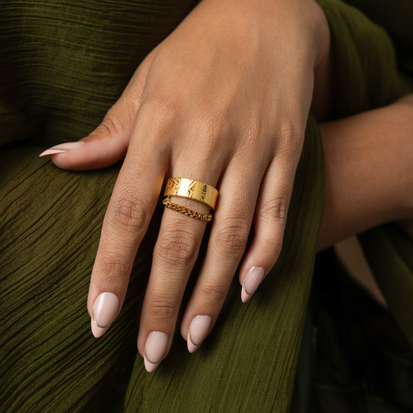 A wide, gold-plated ring with a rectangular face and intricate, abstract designs engraved across the surface. A delicate gold chain is attached to either side of the ring. The ring is worn on a person's finger with light-colored nail polish. Another delicate gold ring is worn on the same finger below the wider ring. The background is a blurred green garment.