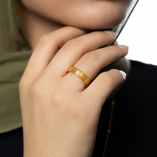 model wearing the gold Be and it is ring, with Arabic engraving visible, hand gently near face, styled with an olive green hijab and black top.