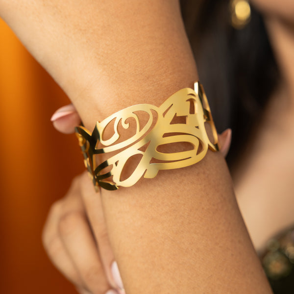 A person's wrist is adorned with a wide, gold cuff bracelet featuring intricate, swirling cut-out designs. The bracelet has an open-ended style, and the delicate patterns create a lacy or filigree appearance.
