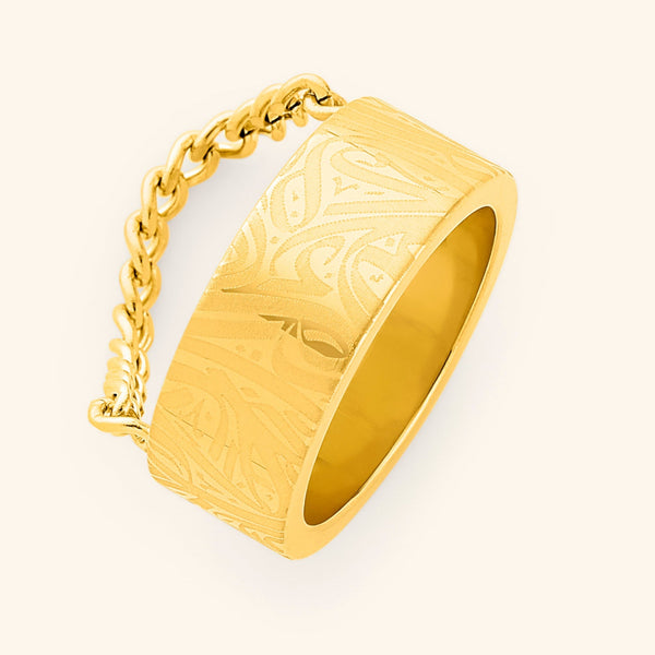 A wide, gold-plated ring with a rectangular face and intricate, abstract designs engraved across the surface. A delicate gold chain is attached to either side of the ring. The ring is displayed against a light beige background.