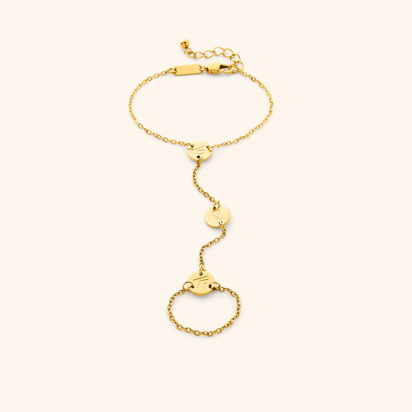 A delicate gold-plated hand chain bracelet featuring a chain that connects a bracelet around the wrist to a ring around a finger, with three small, round charms with intricate cut-out designs along the connecting chain. The hand chain is displayed against a light beige background.