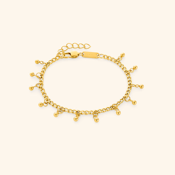 Flat lay of a gold dangling dotted bracelet featuring small dangling beads, displayed against a cream background. The clasp tag shows the engraved text “NOMINAL”.