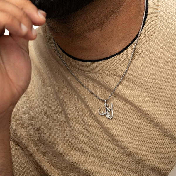 Man wearing a silver 'Faith' calligraphy necklace with a beige shirt, resting hand on chin.
