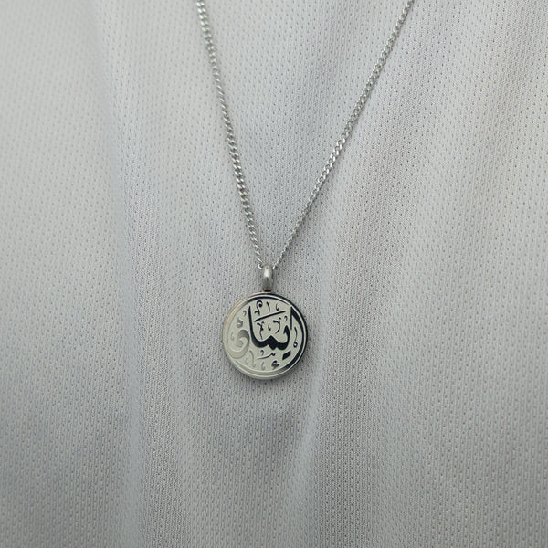 Silver faith token necklace over a white textured shirt, showing the engraved Arabic script “Iman”.
