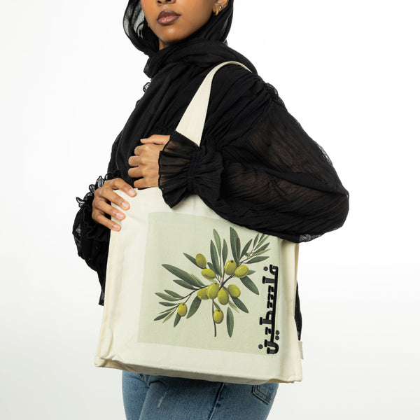 A model holding the Falasteeni Olives Tote over their shoulder, showcasing the tote's size and the prominent olive branch design with the Arabic script for "Palestine."