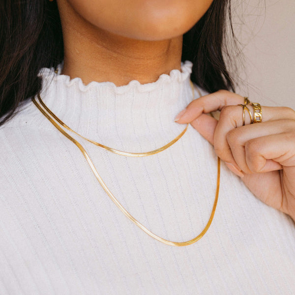 Model wearing two gold herringbone chains over a white ribbed turtleneck, holding one chain with her left hand.
