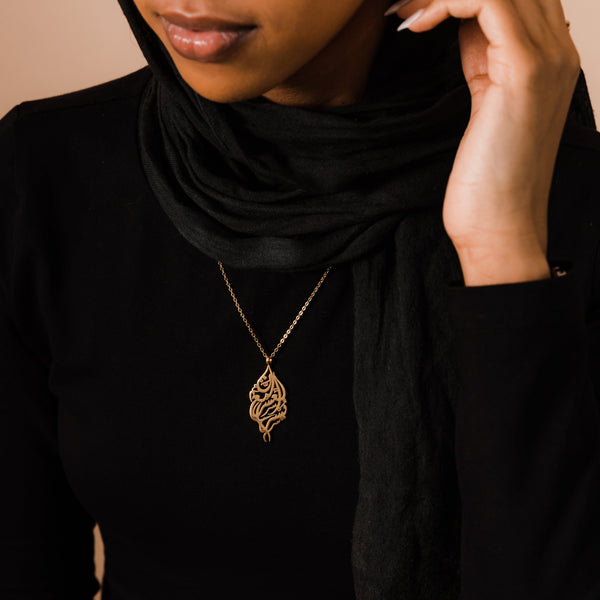A model wearing the 18K gold-plated "Verily, With Hardship Comes Ease" Calligraphy Necklace. The pendant rests gracefully on her collarbone, highlighting the intricate Arabic calligraphy against a neutral-toned outfit.