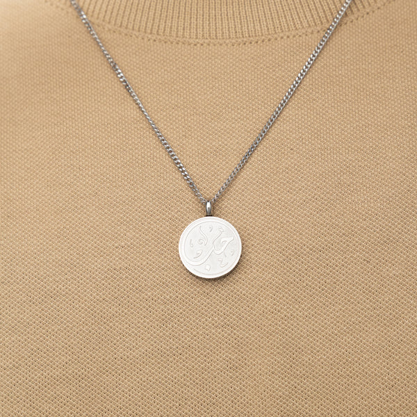 Silver necklace with a circular 'Maktub' calligraphy engraved-pendant worn over a beige knit top.