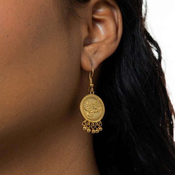 A close-up of a model wearing the gold-plated Maktub earrings. The earrings dangle elegantly, catching the light and highlighting the intricate Arabic engraving and the delicate dangling orbs.
