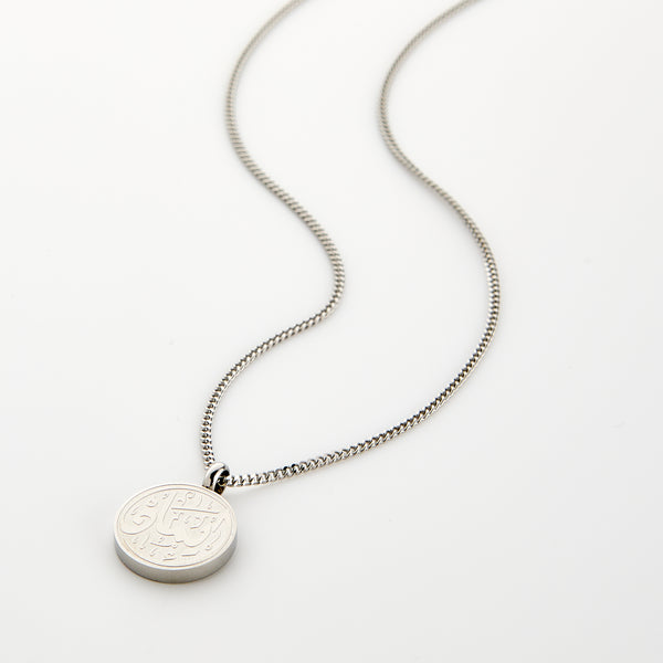 A silver Faith Token necklace with the pendant resting flat on a light background, showing the engraved Arabic script “Iman”.