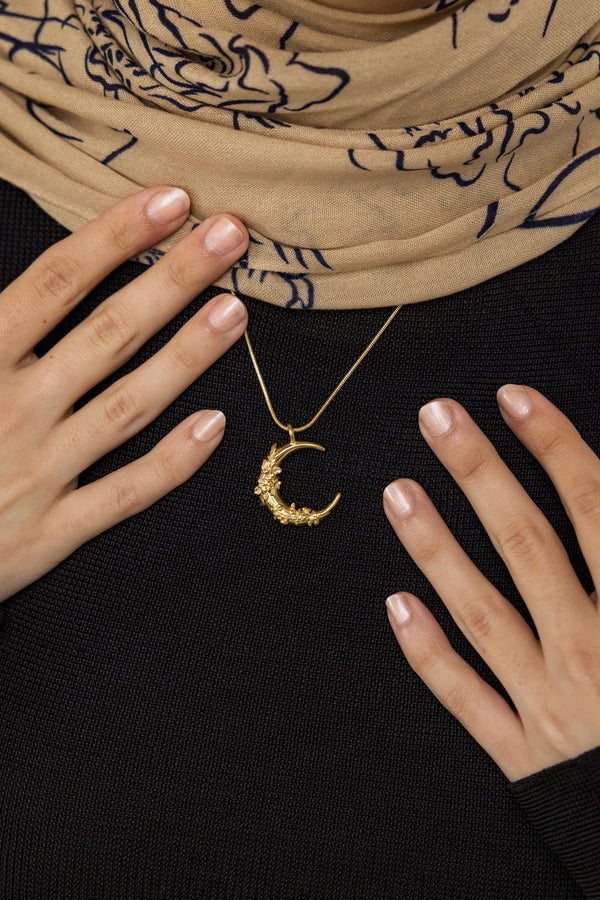 A female model with medium skin tone wearing the 18K gold plated Jasmine Crescent Necklace. The crescent pendant with jasmine vine detailing rests elegantly on her collarbone, complementing her minimalist outfit