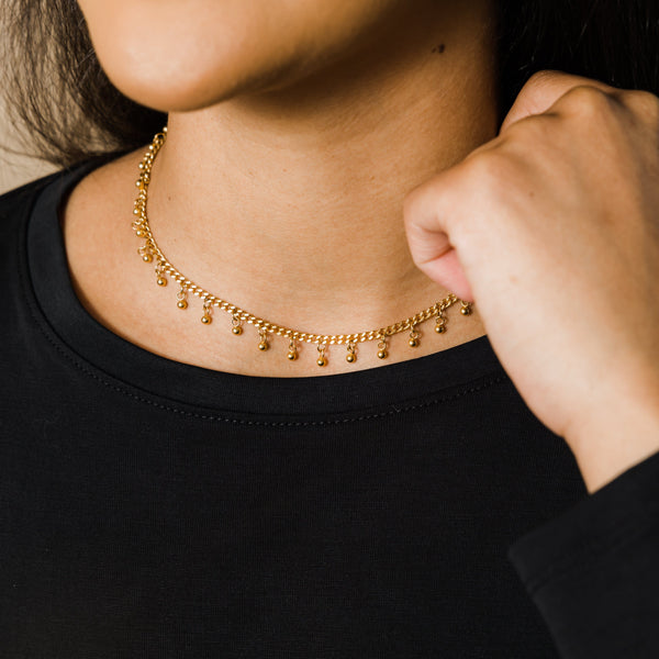 Gold choker with small dangling beads worn by a woman in a black top, hand resting near the neck.