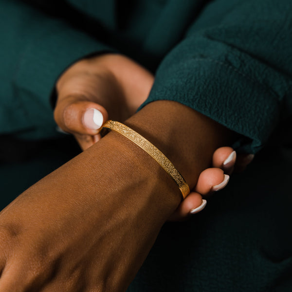 A close-up shot of a person's arm and hand wearing a thin, textured gold bangle bracelet. The person has medium-toned skin and manicured nails with white tips. They are wearing a dark teal, long-sleeved garment. Their other hand is gently touching the bracelet.