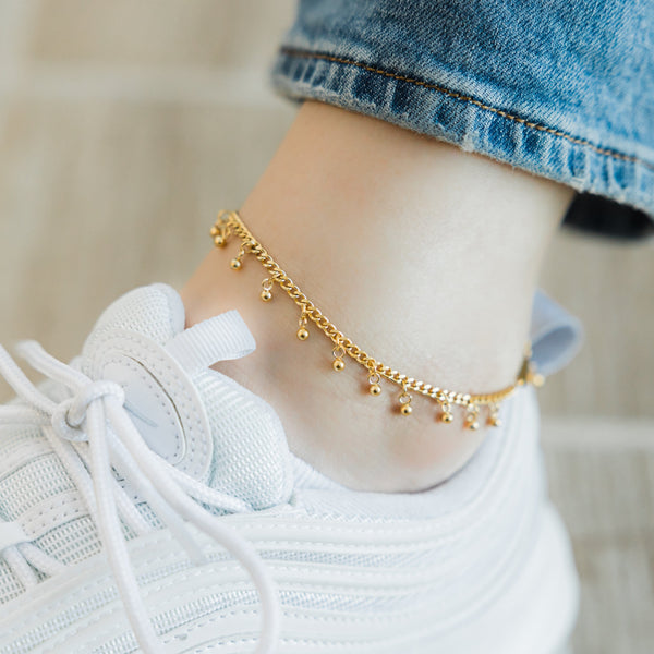 A model wearing the 18K gold-plated Dangling Dotted Anklet. The anklet rests gracefully on her ankle, highlighting the dangling ball-shaped charms against her skin.
