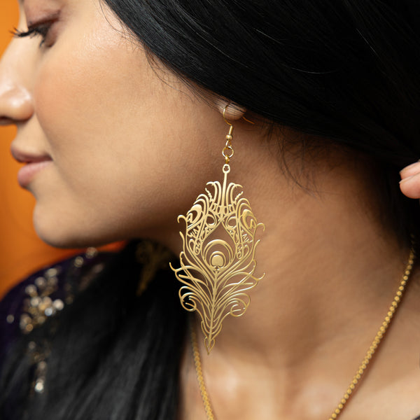 The image shows a person wearing a gold "Peacock Feather Earring" from the Aadil x Nominal collection. The earring is intricately designed in the shape of a peacock feather, with detailed cut-out patterns. The person has dark hair and is wearing a dark blue top with gold embellishments and a gold necklace.