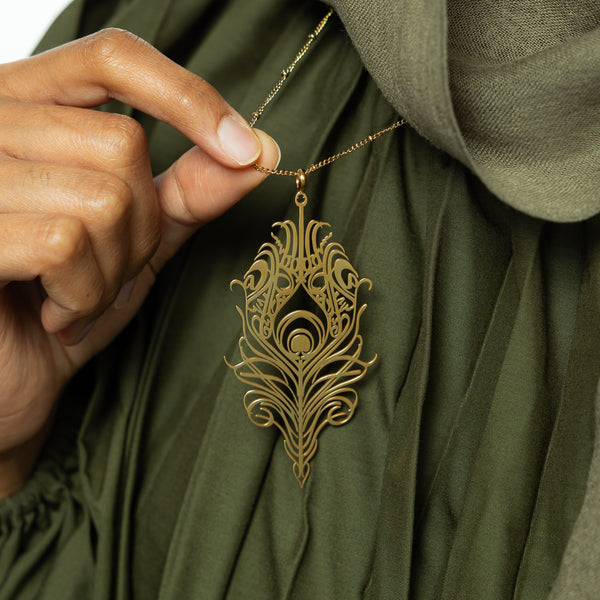 A person's hand is gently touching a delicate gold-plated chain necklace featuring a stylized, peacock feather-shaped pendant with intricate cut-out details. The background is a green garment.