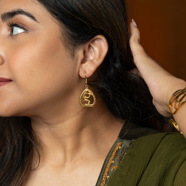 A person wearing a gold "Peepal Earring" from the Aadil x Nominal collection. The earring has a unique, abstract design with swirling lines enclosed within a roughly triangular frame. The metal is a warm gold color. The person is wearing a green top and has a hand near the earring. The image is a product shot, likely showcasing the design and craftsmanship of the earring.