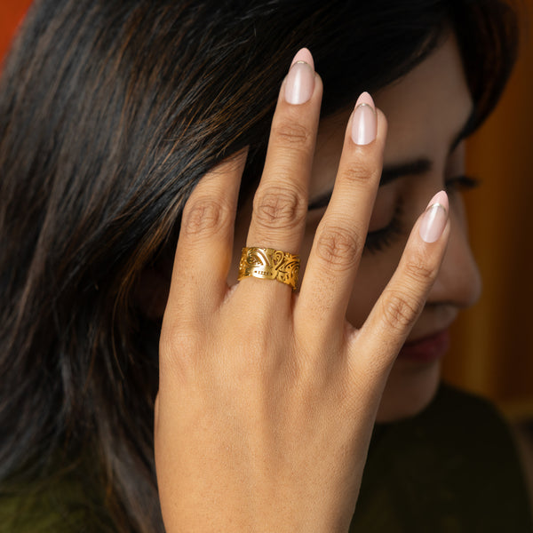 A close-up of a woman with dark hair holding her hand near her face; she wears a wide gold filigree ring on her middle finger and has long, pale pink, almond-shaped nails with metallic tips.