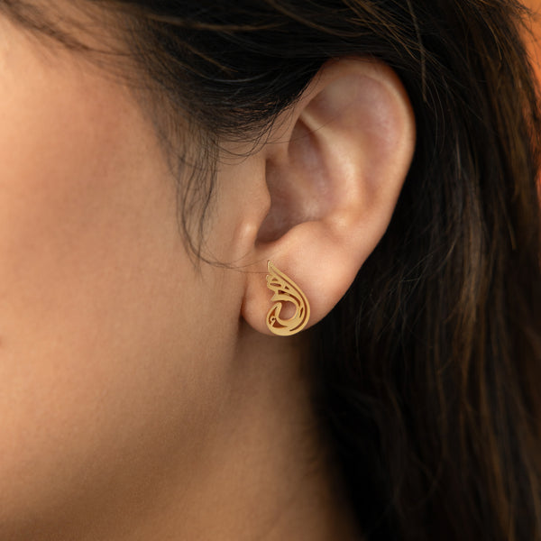 The image shows a person's ear adorned with a gold "Serenity Earring" from the Aadil x Nominal collection. The earring features a unique, abstract, teardrop-like design with swirling cut-out details. The metal has a warm gold color.