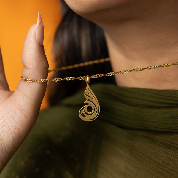 A delicate gold-plated chain necklace featuring a dainty, approximately 2 cm teardrop pendant with intricate, swirling cut-out details and a shiny finish. The necklace is worn by a person with dark hair and is shown against a blurred green garment. A hand with light-colored nail polish is gently holding the chain near the pendant.