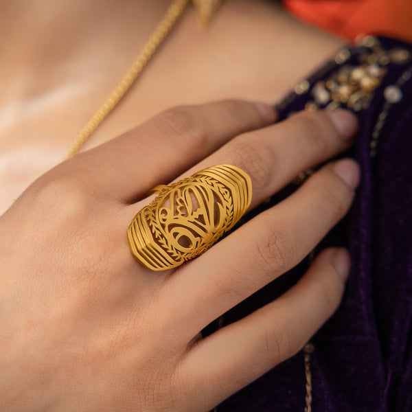 A gold ring with intricate cut-out designs, inspired by ancient artistry, worn on a person's finger. The ring is long and features detailed patterns, possibly with floral or geometric motifs. The person is also wearing a purple top with gold embellishments and a gold necklace. The text "SUFFICIENT (ring) - Women | Aadil x Nominal" suggests this is a product shot for a women's ring, possibly a collaboration between Aadil and Nominal.