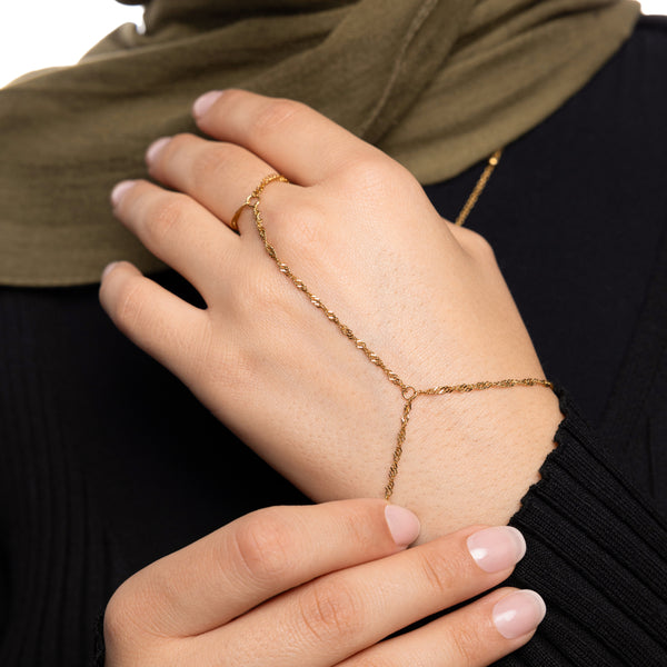 Close-up of a woman’s hand wearing a gold twisted hand chain, styled with a black ribbed top and olive green scarf, showing chain from wrist to middle finger.