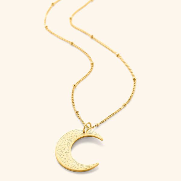 A delicate gold necklace featuring a crescent moon pendant inscribed with Arabic script. The chain is a fine gold link. The necklace is displayed on a light beige background. Given the inscription "With Hardship Comes Ease" and the crescent moon, which holds significance in Islamic culture, it's plausible that the script is related to Islamic teachings and the necklace carries religious or inspirational meaning for the wearer.