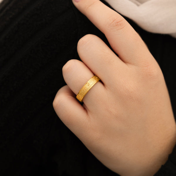 A close-up shot of a person's finger wearing a textured gold ring. The person has light-toned skin and a visible fingernail. The background is blurred and dark, possibly showing a dark garment.