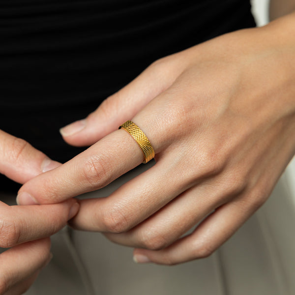 A female model with medium skin tone wearing the 18K gold plated Keffiyeh Ring on her index finger. The ring's intricate keffiyeh-inspired engravings are visible, complementing her minimalist outfit. The model's hand is positioned near her face, drawing attention to the ring's design.