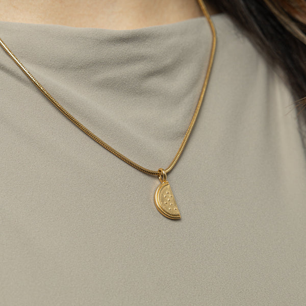 A gold necklace with a pendant shaped like a slice of a watermelon. The watermelon pendant is detailed with small indentations representing seeds. The necklace chain is a thin, flexible gold snake chain. The necklace is displayed against a light beige background.