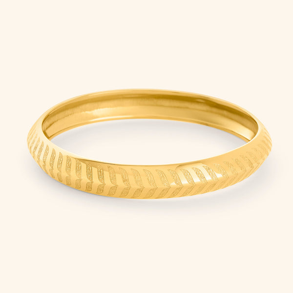 A gold "Chevron Bangle" designed for women as part of the Aadil x Nominal collection by Nominal. 1  The bangle has a smooth, rounded surface and is decorated with intricate, stylized chevron or feather-like carvings that run around its entire circumference