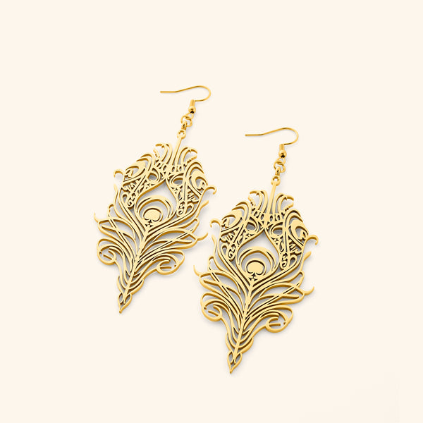 The image shows a pair of gold "Peacock Feather Earrings" from the Aadil x Nominal collection. The earrings are intricately designed in the shape of peacock feathers, with detailed cut-out patterns that mimic the barbs and eye of the feather. They have a warm gold tone and are displayed against a light beige background. This appears to be a product shot showcasing the earrings' design and craftsmanship.