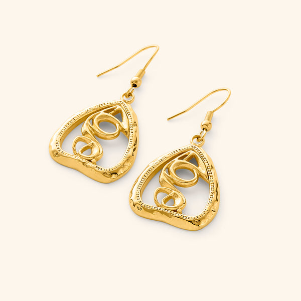 A pair of gold "Hub (Love)" earrings. The earrings feature a unique, abstract design with swirling lines enclosed within a roughly triangular frame. The metal is a warm gold color, and the background is a soft, light beige. The image is a product shot, likely showcasing the design and craftsmanship of the earrings. The text "This image might show: Hub (Love) Earrings" confirms the product name.