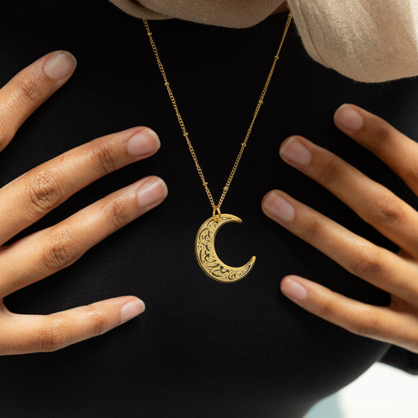 A close-up of a gold necklace with a crescent moon pendant. The pendant is inscribed with Arabic script. The necklace is held up against a blurred background of a person wearing a black top. The focus is on the detail of the pendant and the script. Given the inscription "With Hardship Comes Ease" and the crescent moon, which holds significance in Islamic culture, it's plausible that the script is related to Islamic teachings and the necklace carries religious or inspirational meaning for the wearer.