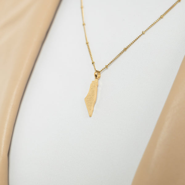A model wearing the 18K gold-plated "With Hardship Comes Ease" Map Necklace. The necklace rests gracefully on her collarbone, highlighting the map pendant with Arabic calligraphy against a neutral-toned outfit.