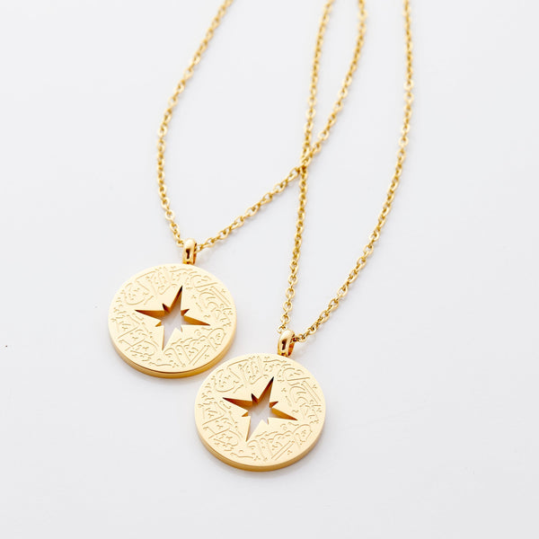 Two matching gold-plated necklaces displayed side by side on a white background. Each features a round pendant engraved with the phrase “Even if I’m not by you, I’m with you” in English and Arabic, accompanied by a compass star symbol.