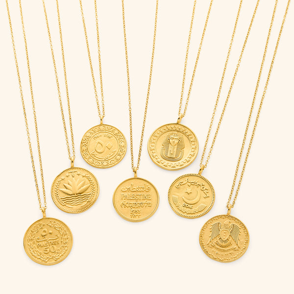 Seven gold-plated country coin necklaces arranged side by side on a neutral background. Each pendant showcases a unique engraved coin design representing a different country, featuring intricate Arabic script, national emblems, and cultural symbols. The 18K gold finish unifies the collection while highlighting the distinct details of each coin.