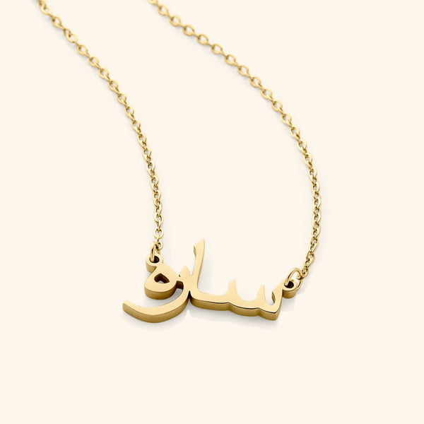 A gold necklace with a name pendant in Arabic script. The chain is delicate and gold. The necklace is displayed on a light beige background.