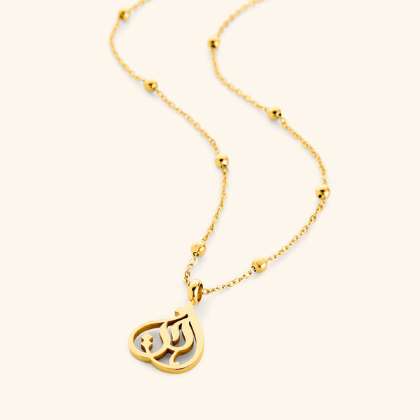 Gold necklace with beaded chain and teardrop-shaped pendant featuring Arabic calligraphy spelling “Sister”, photographed flat against a cream background.