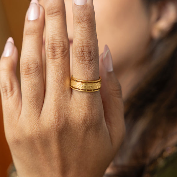 A close-up shows a person's hand with manicured nails wearing a gold ring with intricate, woven designs on their ring finger. The ring features detailed patterns and a subtle texture. The person is wearing a dark top with a floral pattern. The background is slightly blurred.