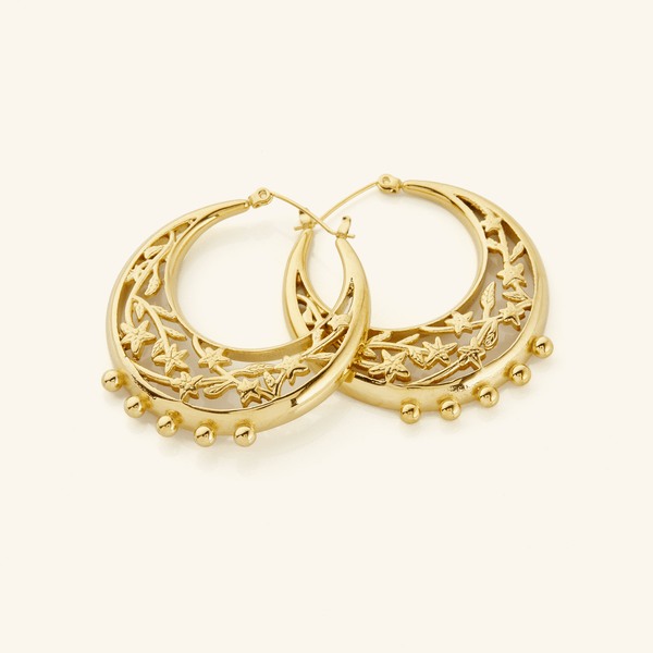 A close-up of the Jasmine Crescent Hoop Earrings in 18K gold plating. The earrings feature a chunky hoop design with an intricate jasmine vine elegantly intertwining within the crescent outline, creating a harmonious blend of bold design and delicate detailing