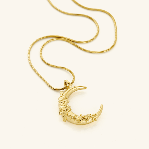 A close-up of the Jasmine Crescent Necklace in 18K gold plating, featuring a crescent-shaped pendant intricately adorned with a jasmine vine design, suspended from the Harmony Chain.